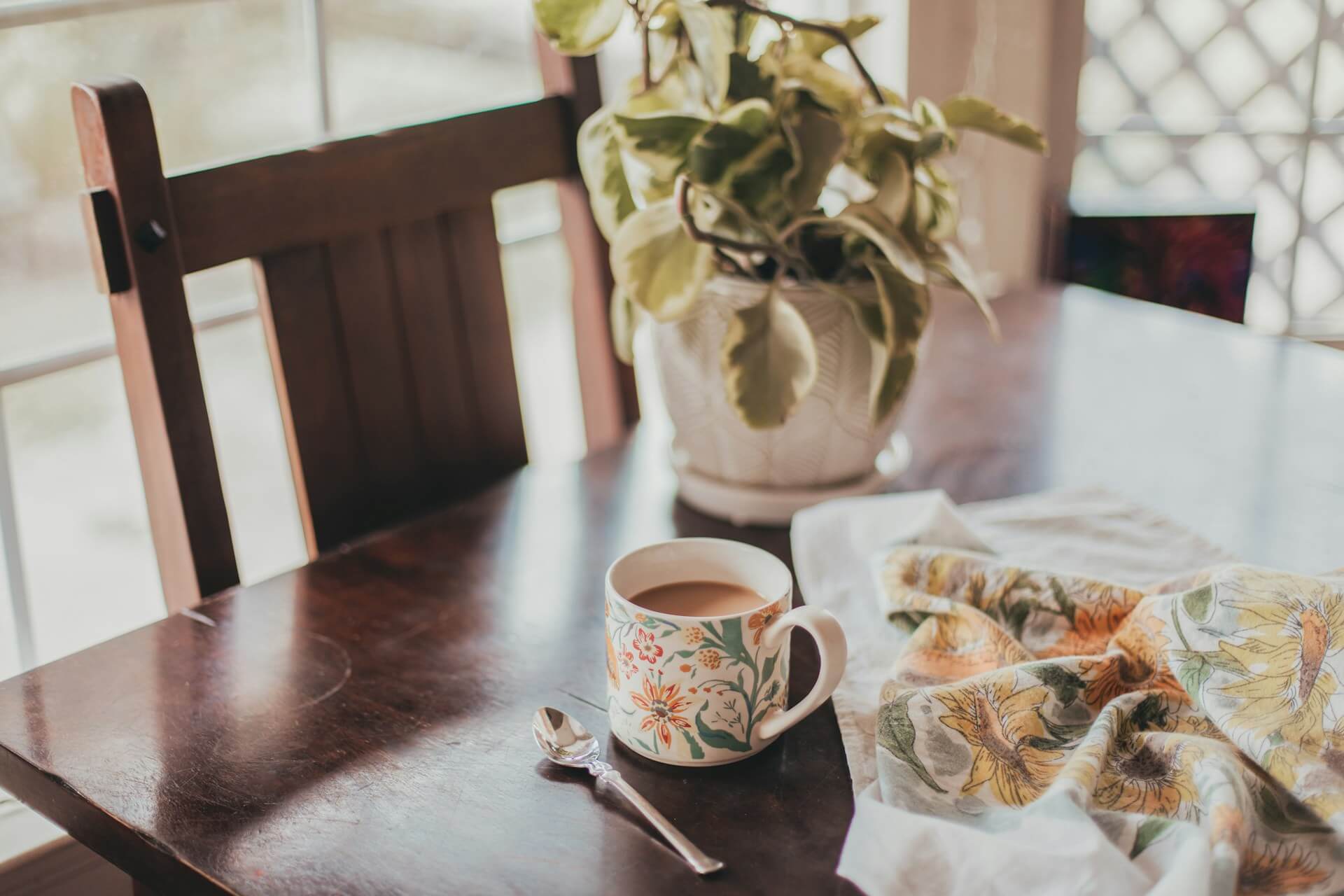A cup of tea with a teaspoon and tea towel on a wooden table, beside a small potted plant.